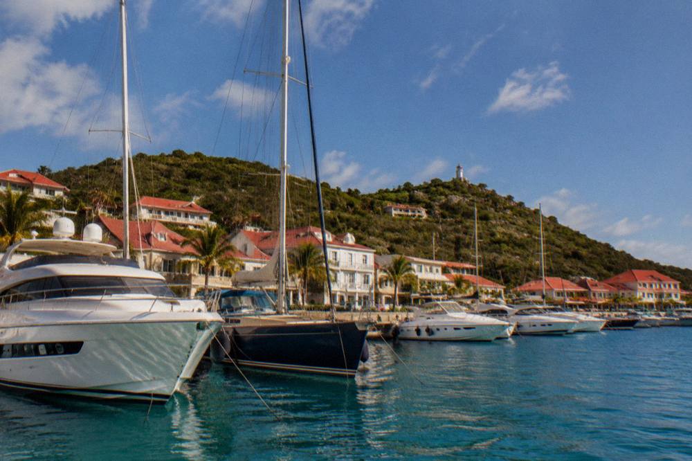 A row of yachts moored along a harbor quay with white buildings behind