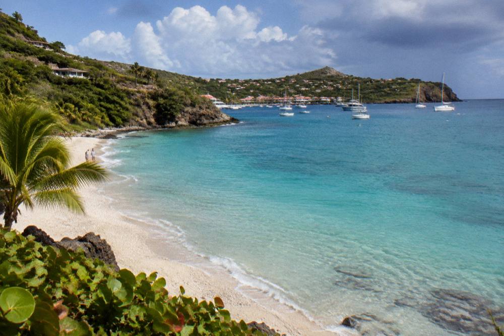 An empty white-sand beach with gentle turquoise water and green headlands in the distance