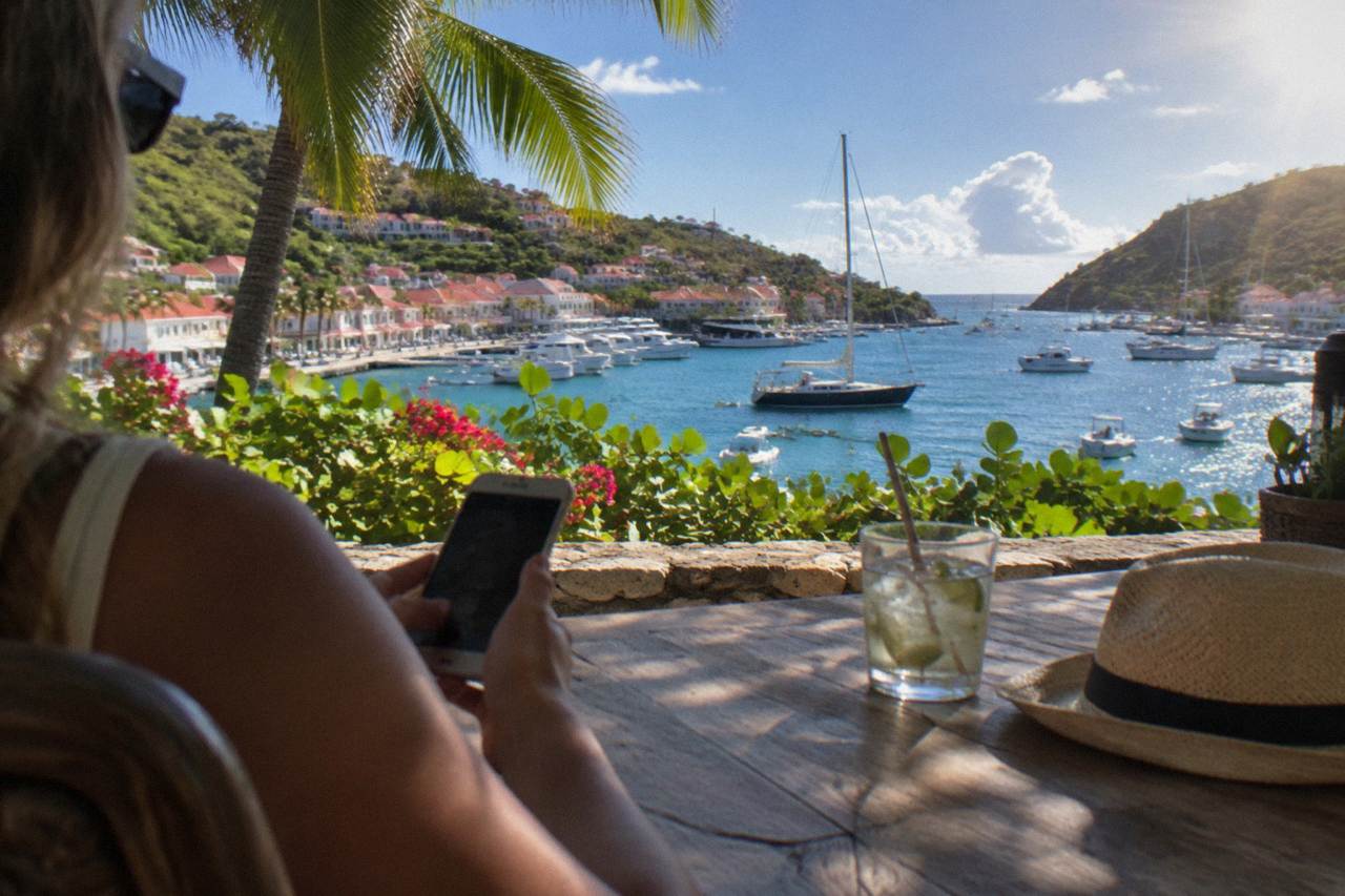 "A visitor using a phone at a shaded table above a Caribbean harbour with white boats