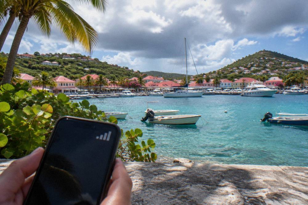 Hands holding a phone showing a signal bar icon beside a departure board at a small Caribbean airport