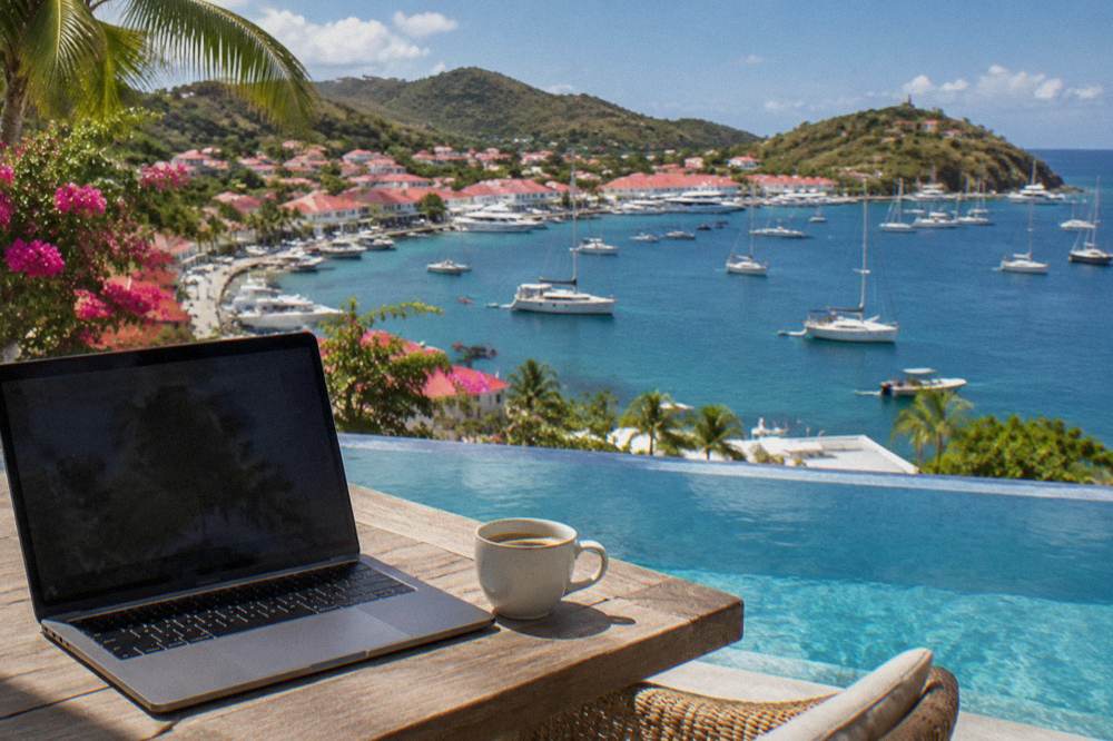 A laptop and coffee on a wooden table beside an infinity pool overlooking a bay with sailboats