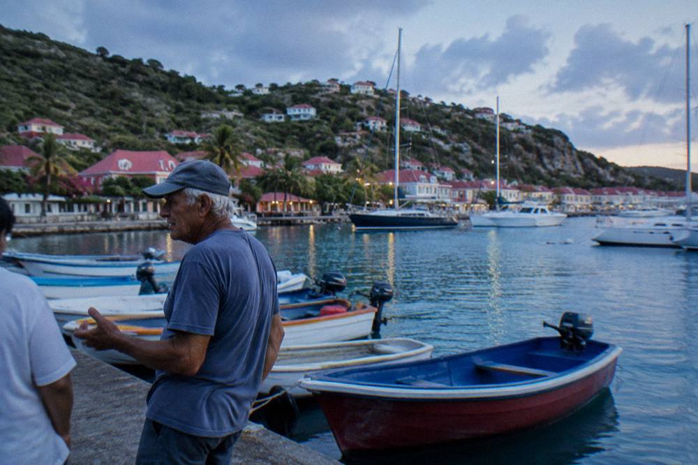 An older fisherman speaking beside small colourful boats at a quiet Caribbean quay