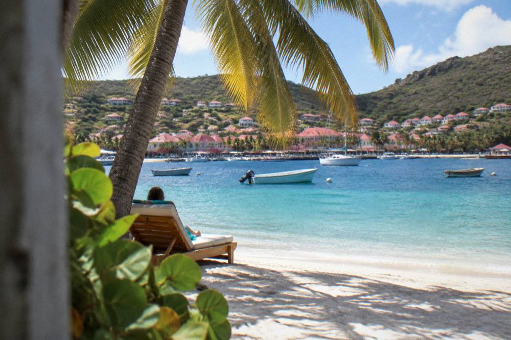 Sun lounger under a palm tree on white sand with clear shallow water beyond