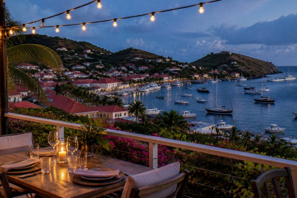 Evening table setting on a terrace with string lights above a harbour with boats at anchor