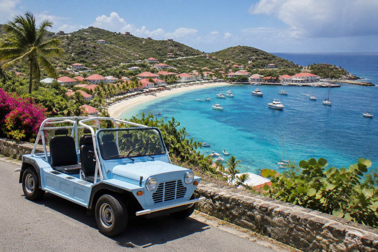 An open-air Mini Moke parked beside a narrow hillside road overlooking a turquoise Caribbean bay