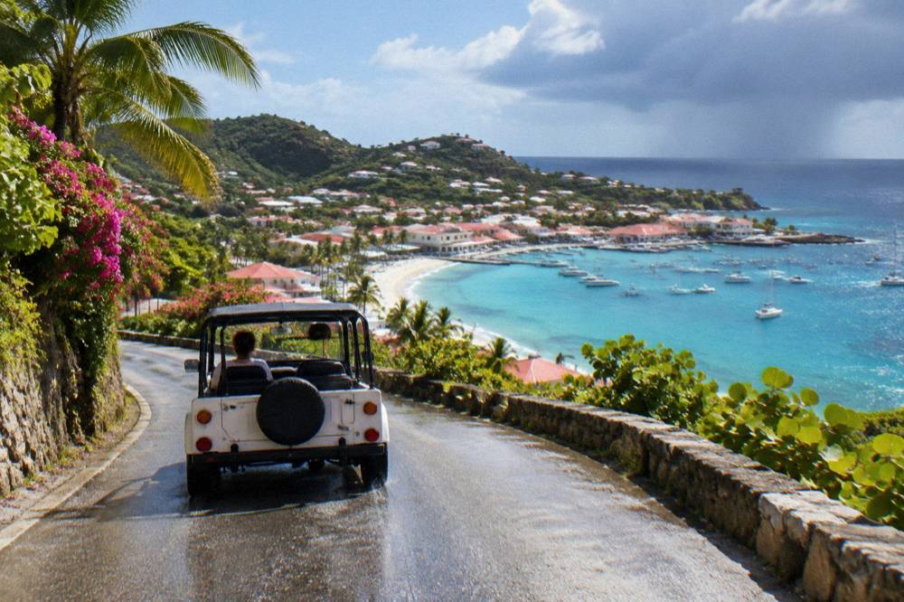 A Mini Moke driving a steep winding road with green hills on one side and sea view on the other
