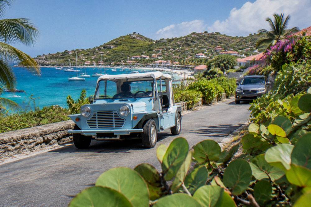 A Mini Moke meeting another car on a narrow island road lined with low tropical vegetation