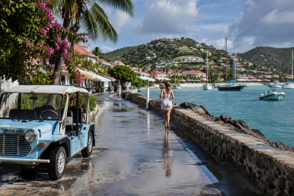 A Mini Moke parked near a harbour wall with sailboats and yachts moored beyond