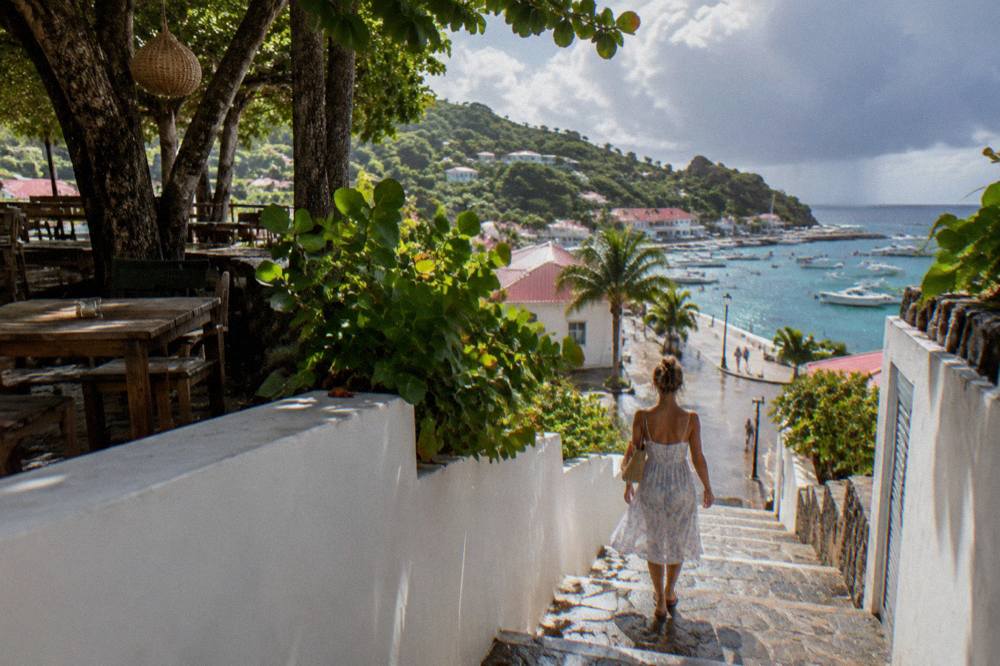 A woman in a light summer dress walking a stone staircase between white walls toward a lit harbor street