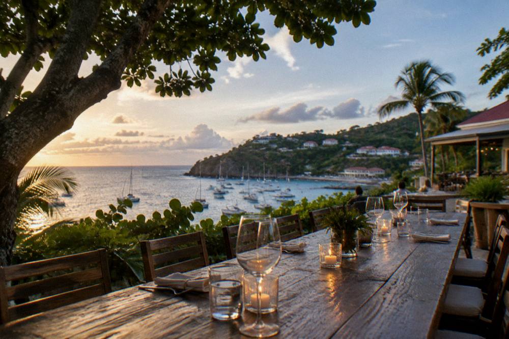 A long wooden restaurant table on a terrace with wine glasses and candles lit against a dark sea horizon