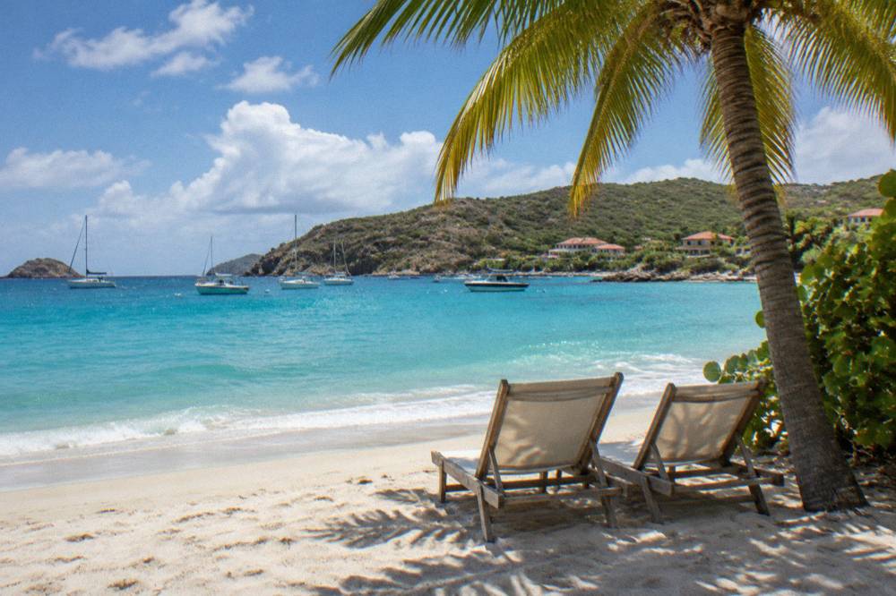 Two empty beach chairs under a palm tree beside calm turquoise water