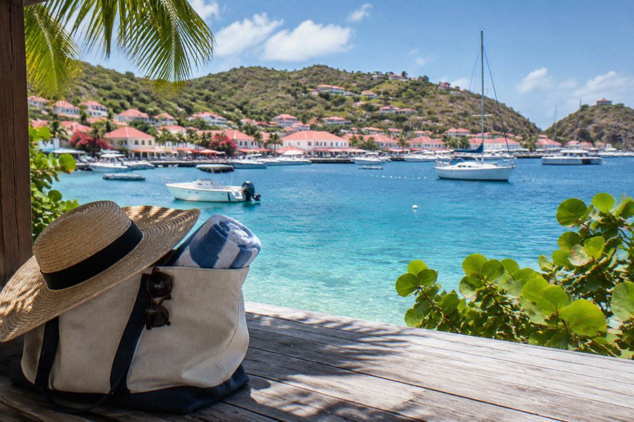 A canvas beach bag with a sun hat, sunglasses and a rolled towel on a wooden deck overlooking turquoise Caribbean water