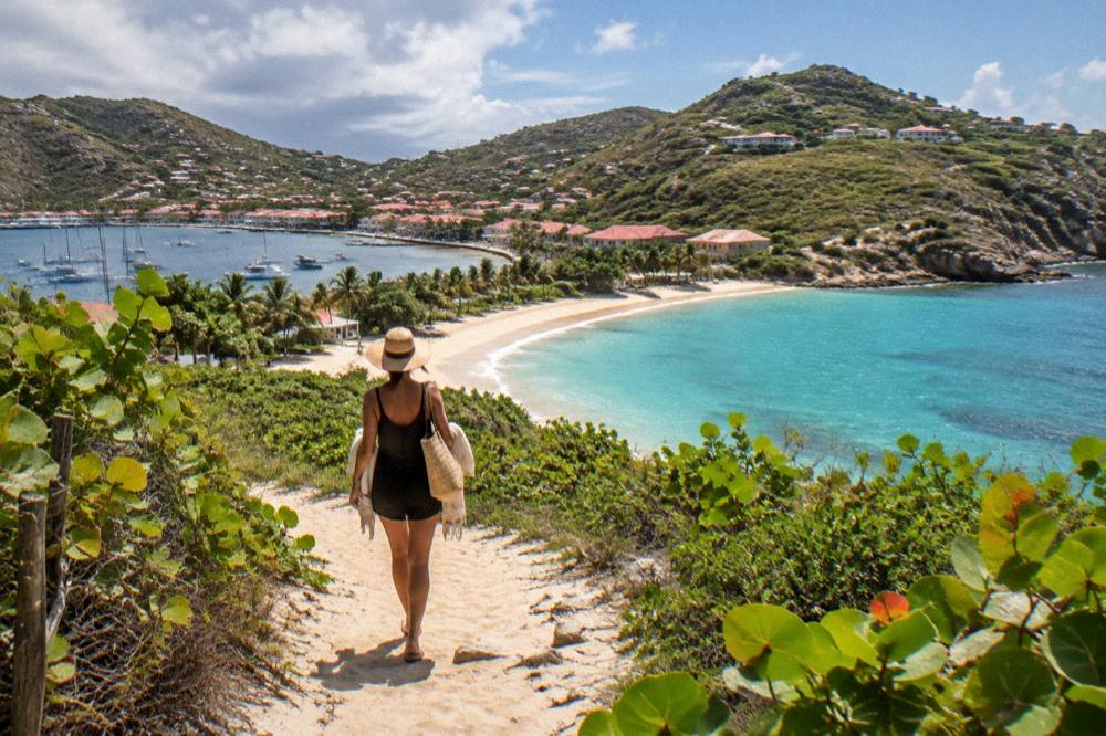 A traveler walking away along a sandy path toward a crescent beach with turquoise water and green headlands