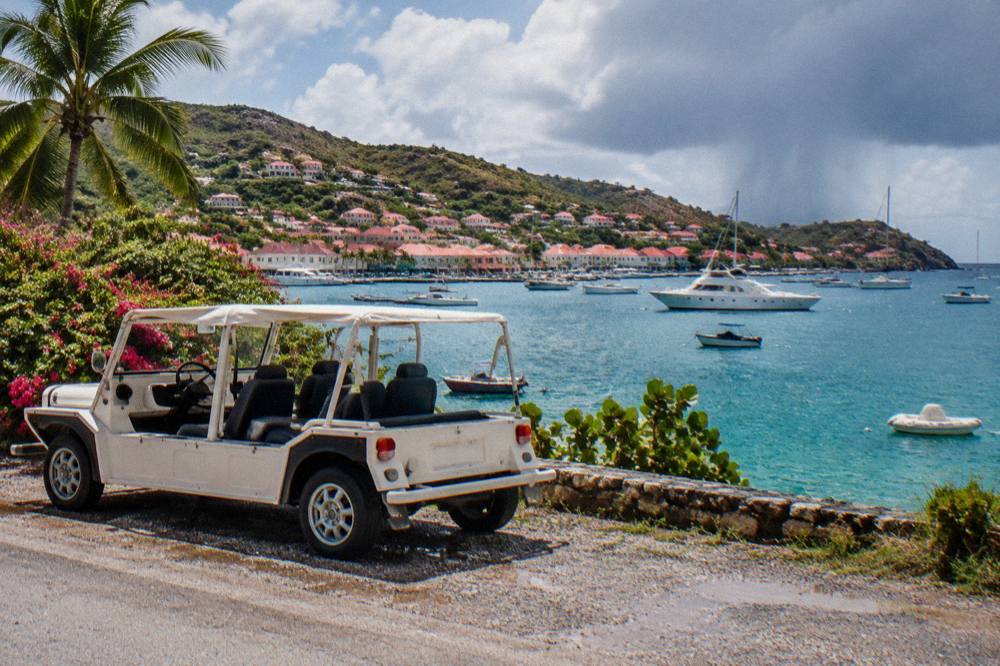 A white Mini Moke parked on a gravel shoulder beside a coastal road on St Barts