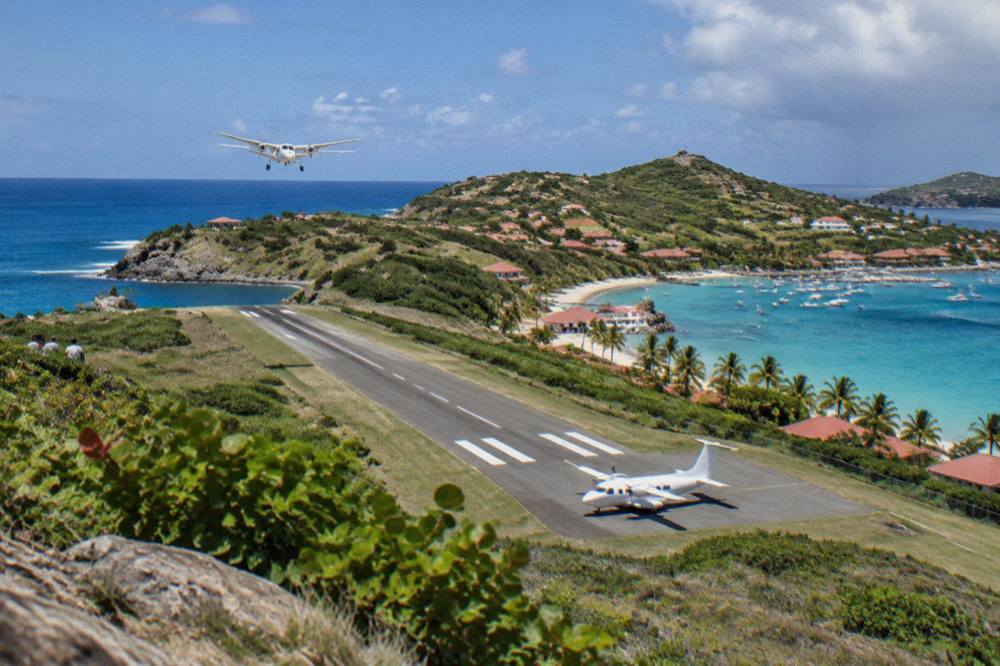 A small white turboprop aircraft parked on a short runway between green hills beside the sea
