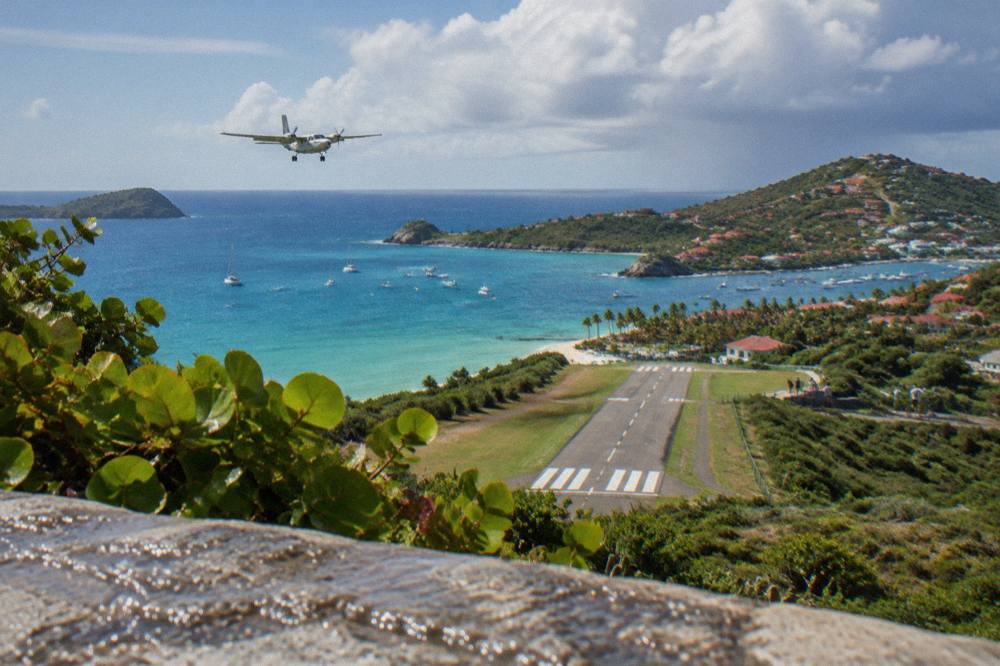 A steep aerial view of a Caribbean hillside runway with turquoise bay water beyond