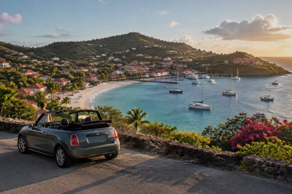 A small open-top car parked at a viewpoint overlooking a Caribbean bay with sailboats
