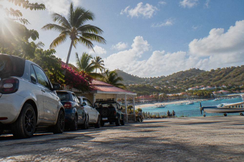 A row of small rental cars and an open-top vehicle parked near a small Caribbean airport building