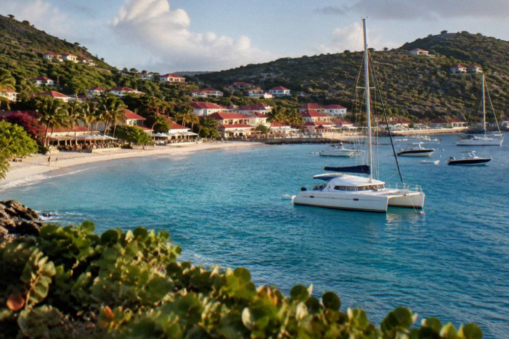 A catamaran anchored in a clear bay with a wooded hill and small houses above the shore