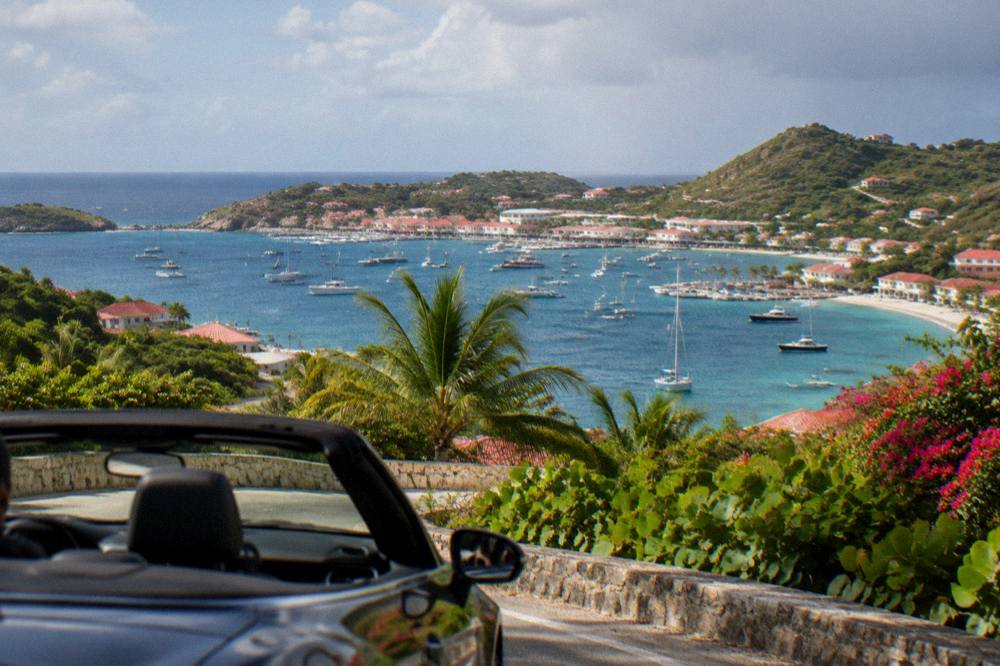 An open-top car on a hill road at golden hour with a harbour town and yachts below