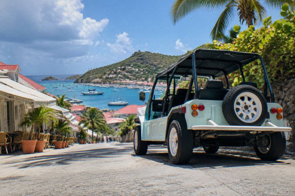 An open-air Mini Moke parked on a hillside road above the sea