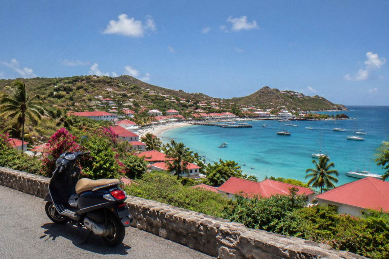 A scooter parked on a narrow hillside road overlooking a Caribbean bay with red rooftops