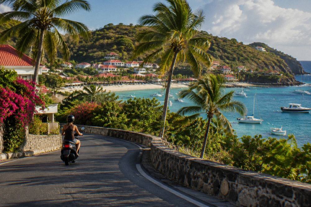 A rider on a scooter rounding a curve with palm trees and the sea beyond
