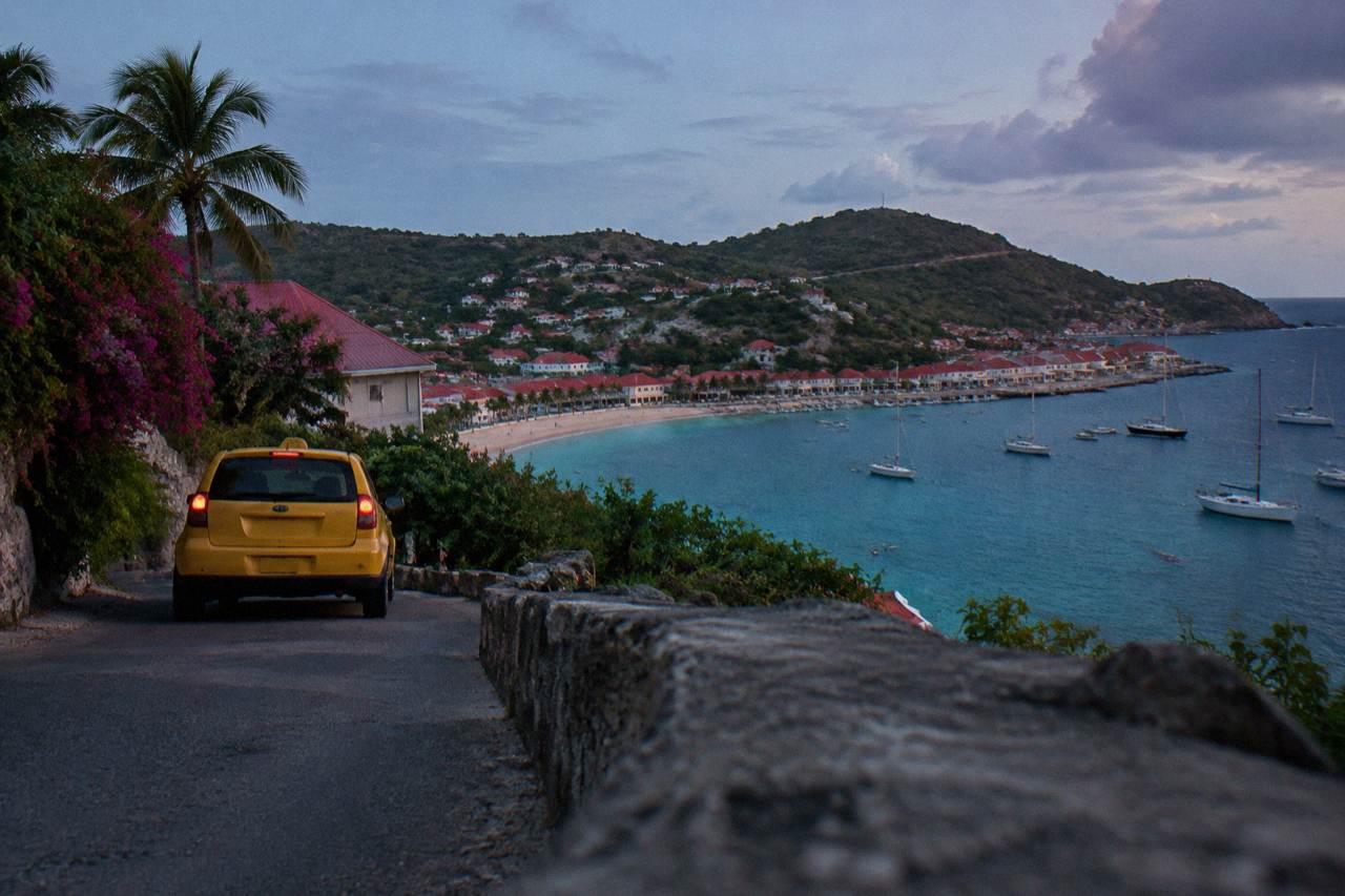 A small taxi waiting on a narrow hillside road above a Caribbean harbor with red roofs