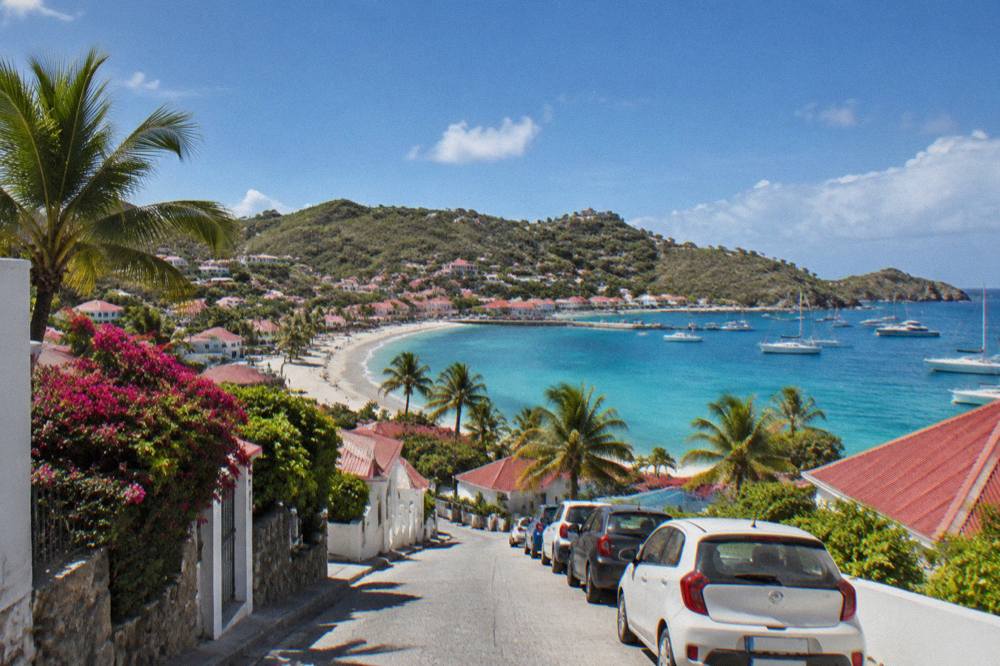 A row of small cars parked on a steep narrow street with white buildings and palm trees