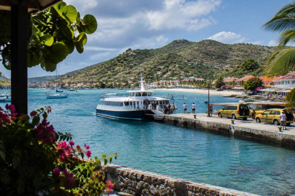 A passenger ferry docking at a small Caribbean quay with taxis waiting on the waterfront road