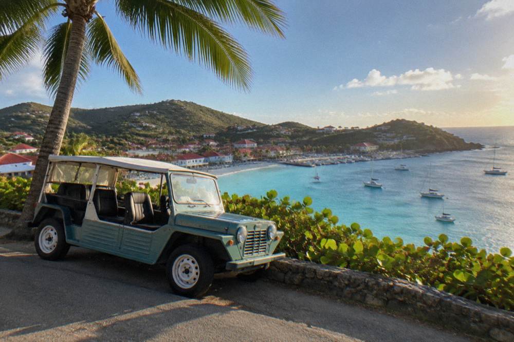 An open-sided Mini Moke parked beside a palm tree on a hillside road above turquoise water