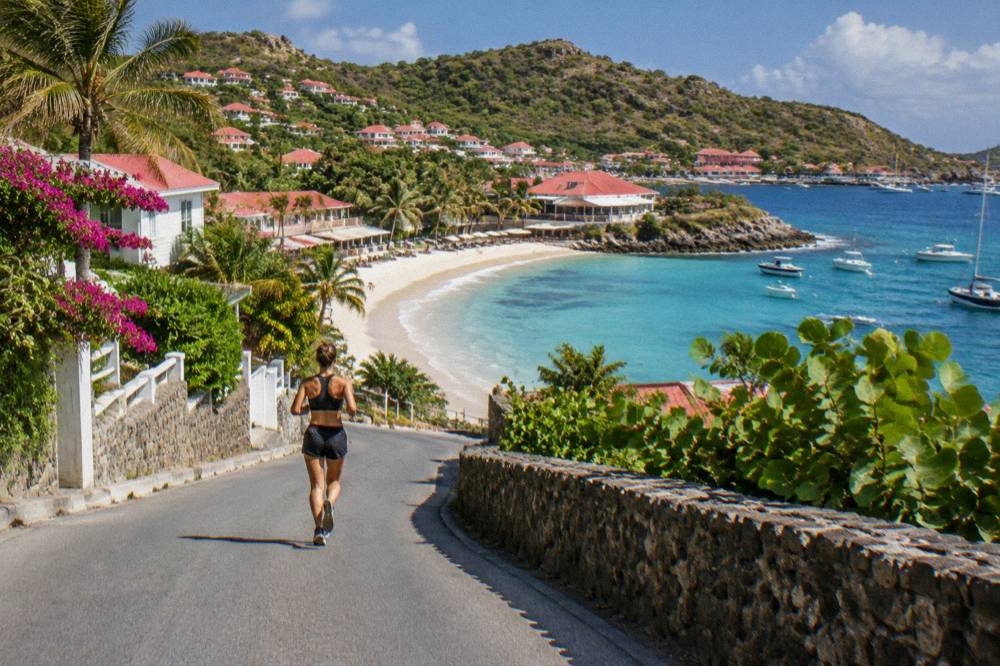 A steep coastal road curving down toward a sandy beach and rocky headland in bright sunlight