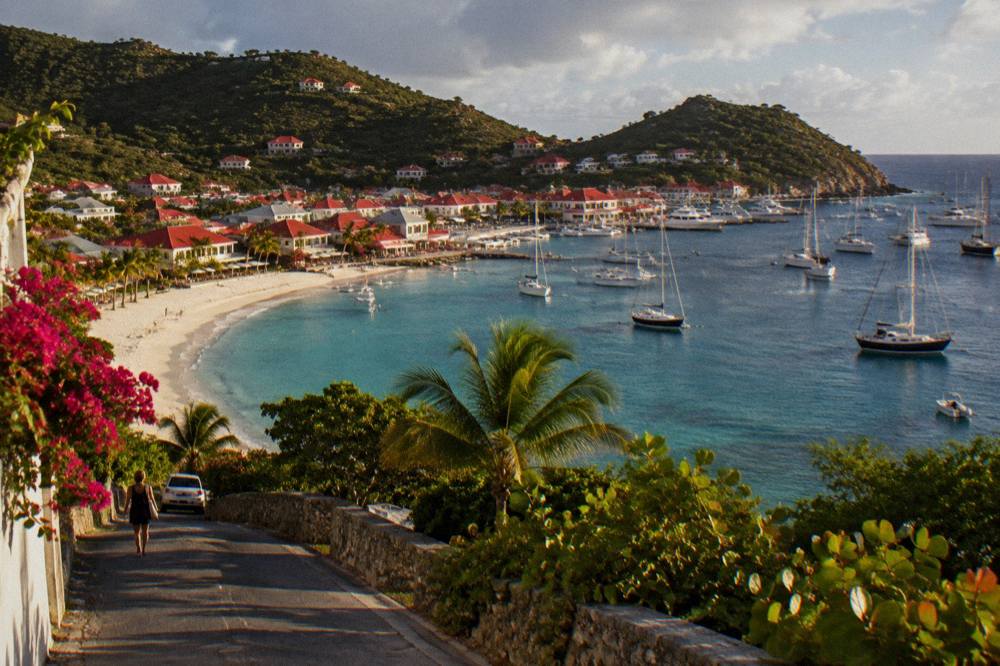 A harbor town at dusk filled with masts and lit waterfront buildings seen from a hillside road