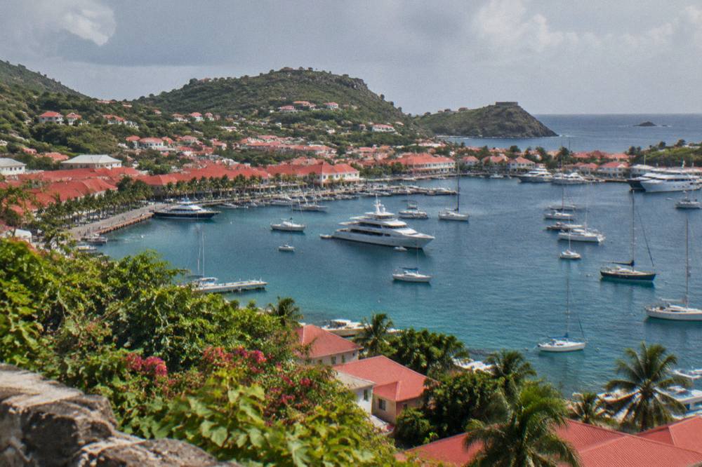 A red-roofed Caribbean harbour town seen from a hillside above moored yachts