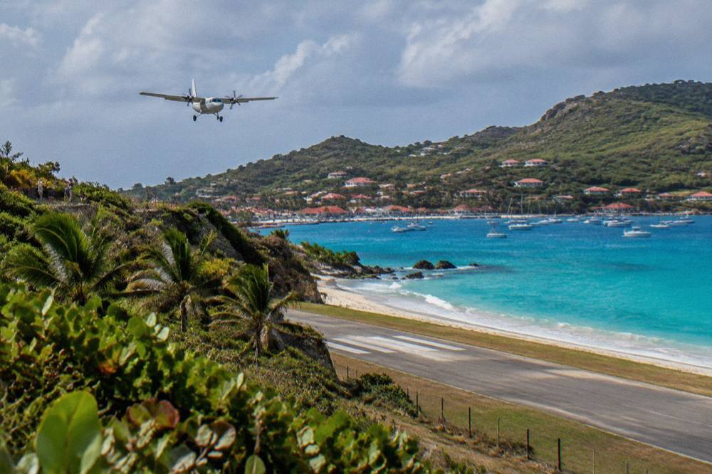 Small turboprop aircraft on a short runway beside a Caribbean beach with hills behind