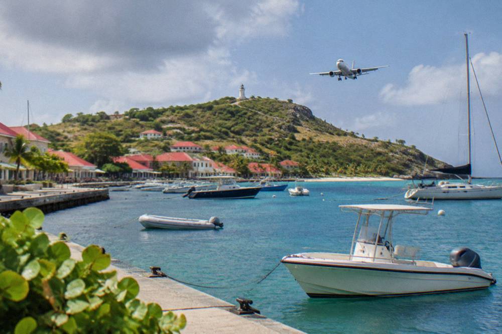 Aircraft on final approach over a green hill toward a beachfront runway in the Caribbean