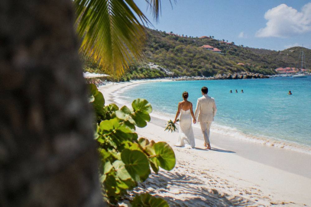 A bride and groom walking on a white sand beach with palm trees and clear shallow water