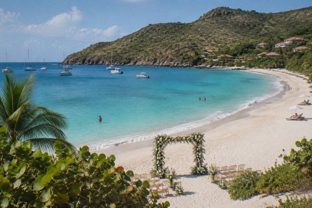 A wedding arch of white flowers on sand with turquoise water and a headland behind