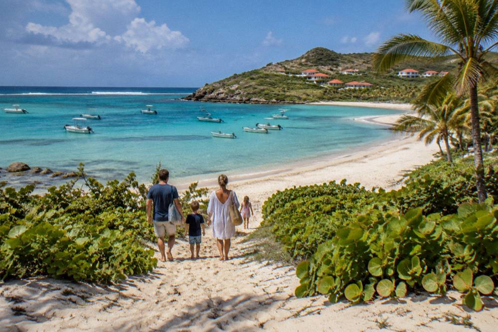 A family walking down a sandy path toward a wide empty Caribbean beach lined with low green vegetation