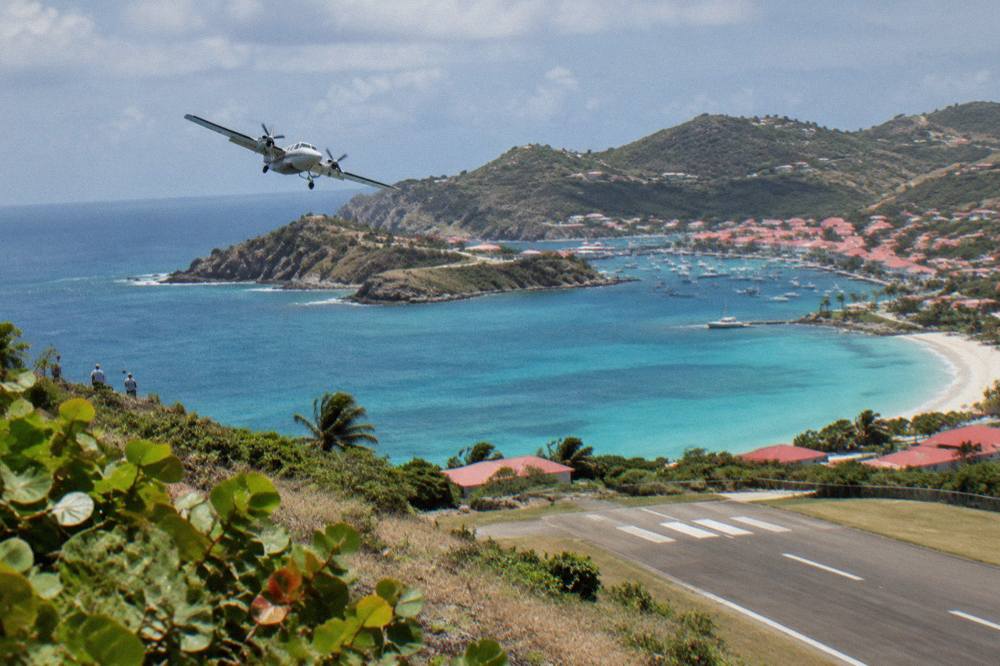 A turboprop plane banking over turquoise Caribbean water toward a hilly island
