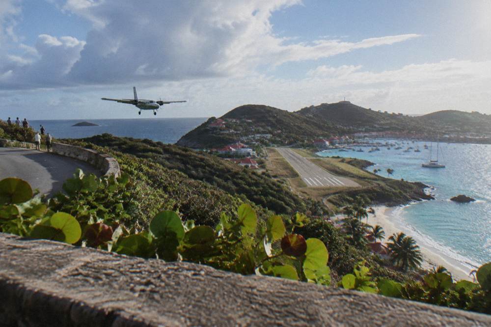 A steep aerial view over a hilltop road toward a short runway beside a harbor town
