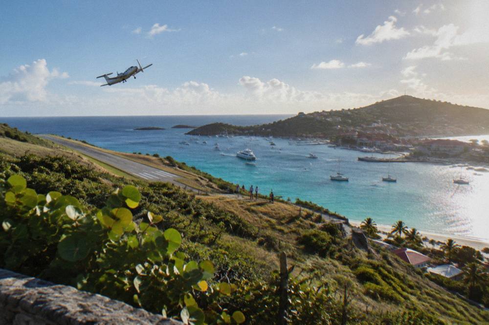 A twin-engine turboprop climbing steeply over turquoise Caribbean water shortly after takeoff