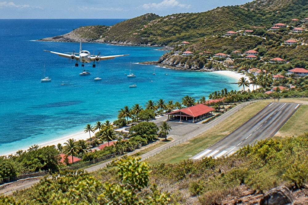 A small terminal building with a red roof beside a short runway and palm trees on a Caribbean hillside