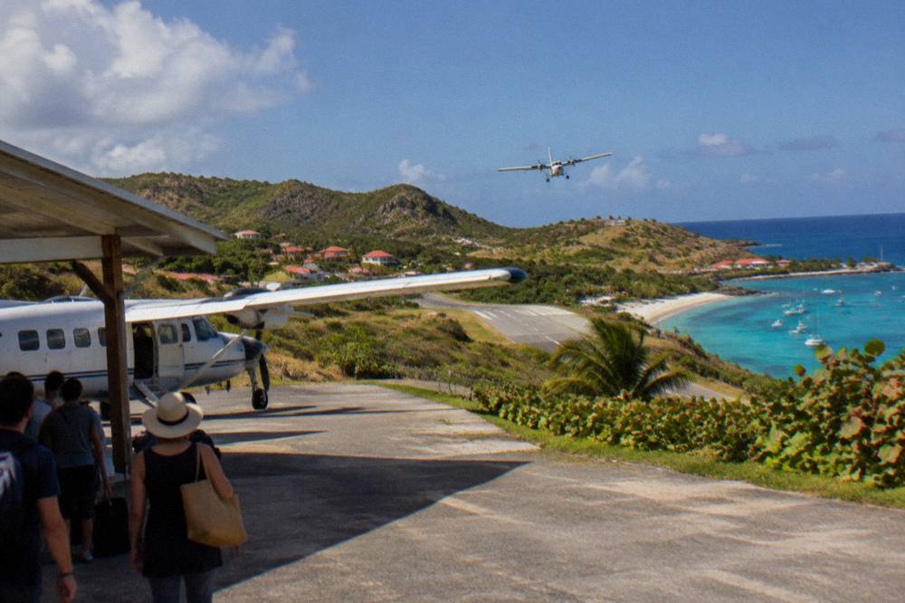 An open-sided small aircraft parked at a Caribbean terminal with passengers waiting in shade nearby
