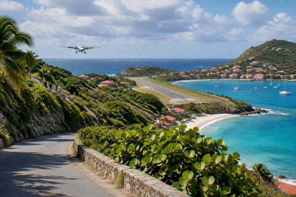 A steep green hillside road descending toward a small coastal airstrip and turquoise bay