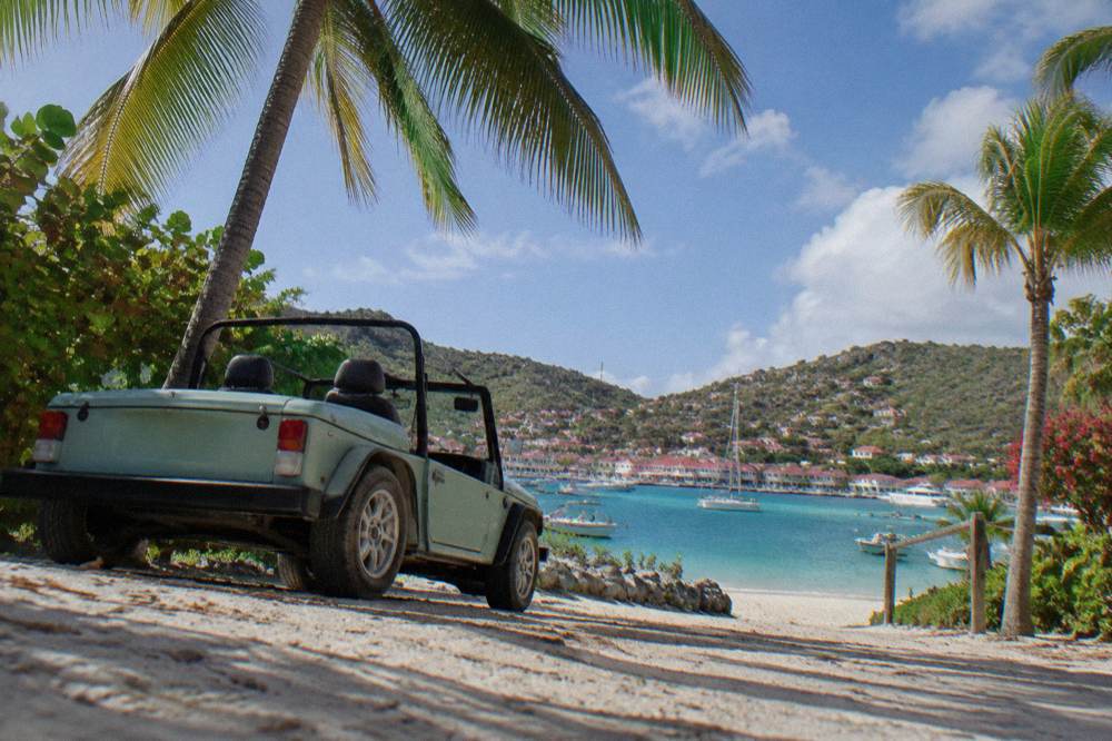A small open-top rental car parked beside palm trees near a beach entrance on St Barts