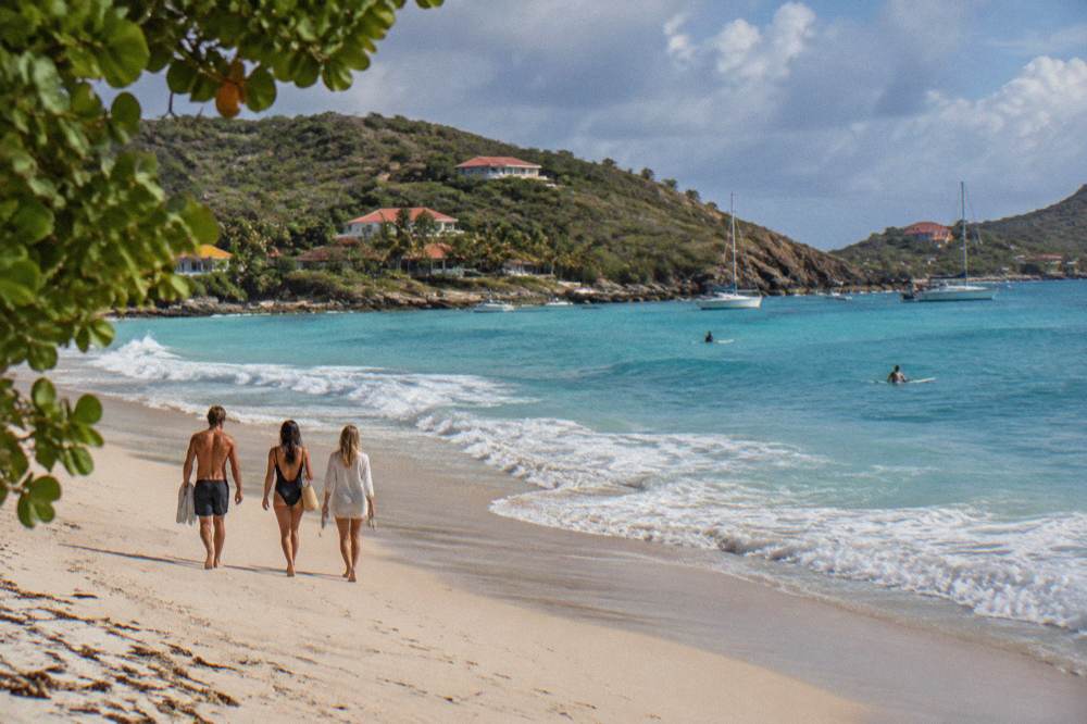 Friends walking barefoot on wide white sand toward turquoise water