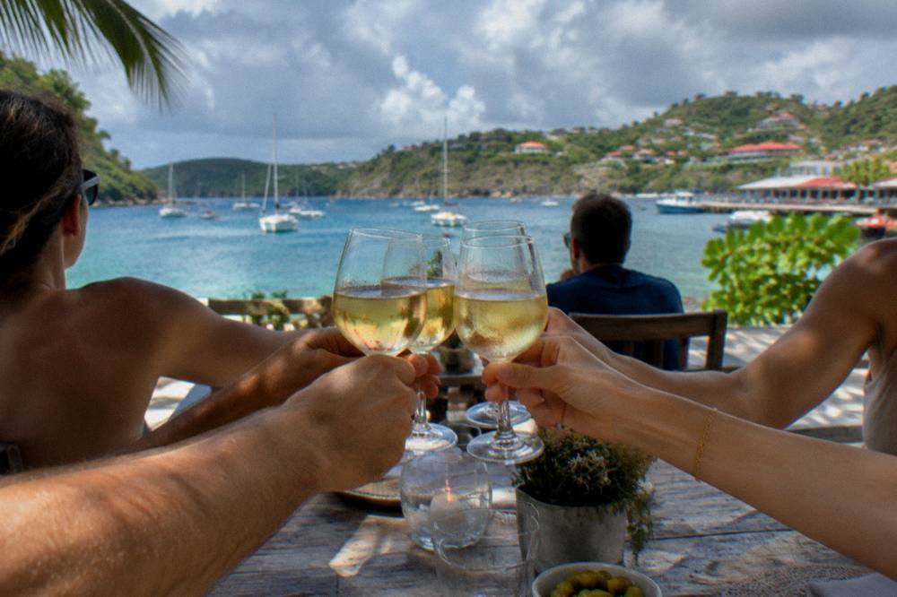 Friends toasting with glasses at an outdoor table as evening light falls on a harbour