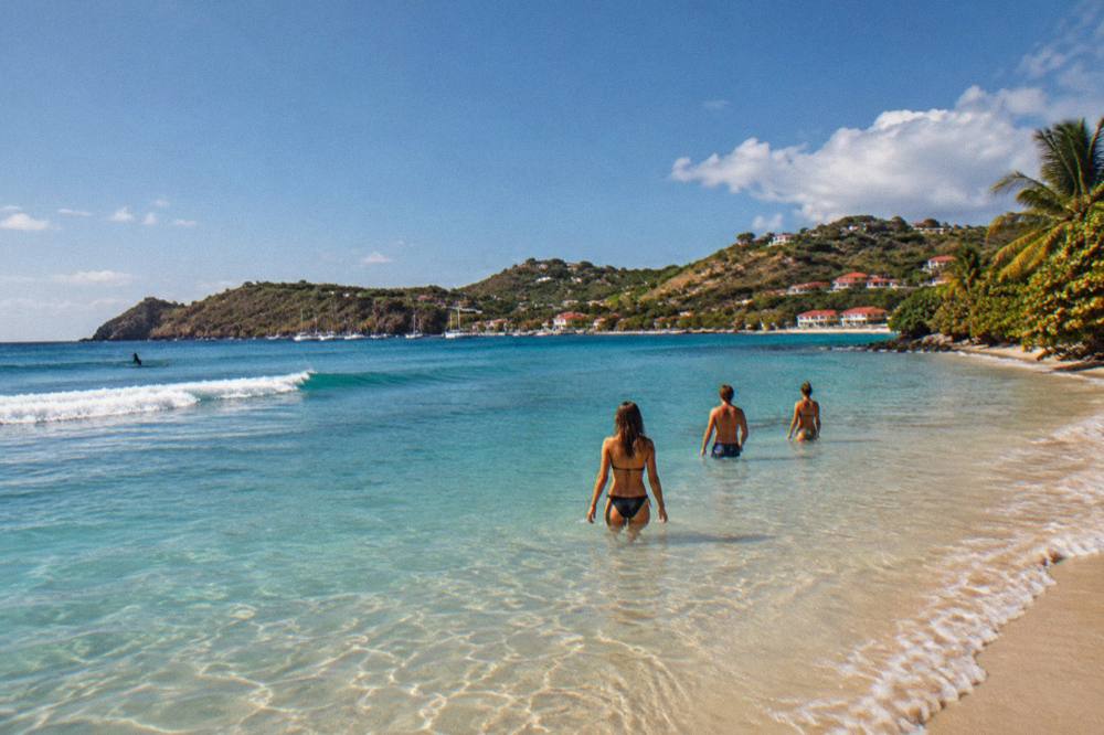 Friends in swimwear wading into clear shallow water under a bright sky