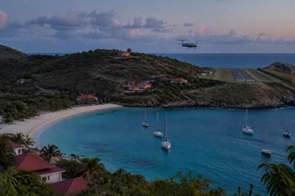 A small turboprop aircraft flying low over a hill toward a short Caribbean runway